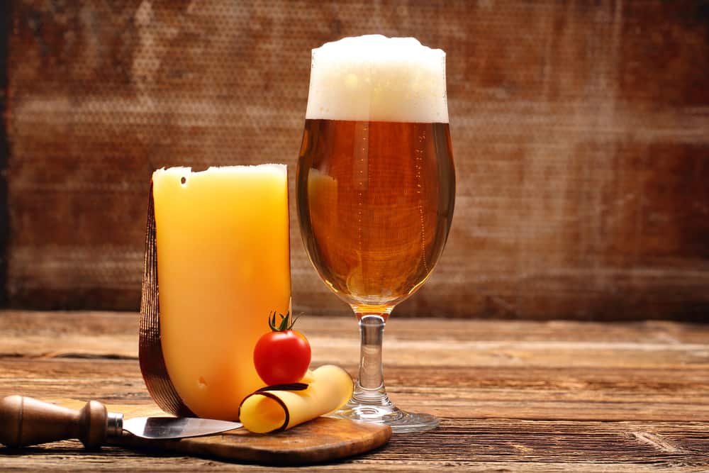 Et glas øl med skum ved siden af en gul ost på højkant - med osteskærer ved siden af. Alt placeret på et træbord.